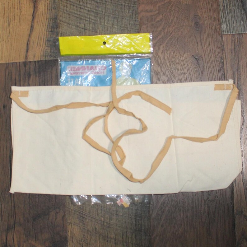 May include: A cream-coloured apron with tan ties, lying on a wooden surface. A package with the text "EMMA" is visible above the apron. The apron is made of a plain, unadorned fabric.