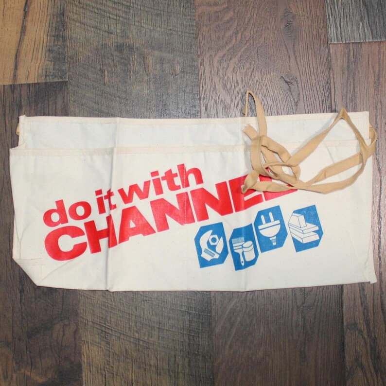 May include: Cream canvas apron featuring the words "do it with CHANNEL" in red, and blue tool icons. The apron has multiple pockets and a tan tie. The apron is on a wooden surface.
