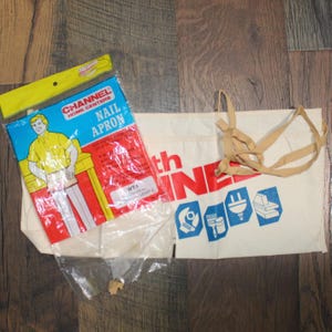 May include: A nail apron and a canvas tote bag are shown on a wooden surface. The nail apron is in a plastic package with the text "CHANNEL HOME CENTERS NAIL APRON". The tote bag has the text "th LINE" and tool graphics.