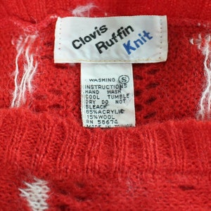 Pode incluir: Uma etiqueta branca com texto vermelho e azul que diz "Clavis Ruffin Knit" e instruções de lavagem. A etiqueta está presa a um suéter de tricô vermelho com um padrão branco.