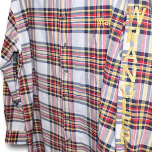Peut inclure: Une chemise à carreaux à manches longues avec un fond blanc et un motif à carreaux rouge, jaune et bleu. La chemise a un devant boutonné et un logo Wrangler sur la poitrine gauche. La manche droite a le mot "WRANGLER" brodé en or.