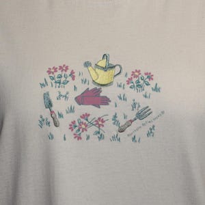 May include: Cream-colored sweatshirt featuring a garden-themed design. The graphic includes a yellow watering can, pink gloves, a trowel, a garden fork, and pink flowers. The text "Northern Reflections" is printed on the bottom right.
