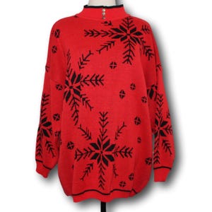 Pode incluir: Camisola vermelha com um padrão de floco de neve preto. A camisola tem gola alta e fecho de correr.