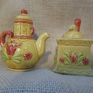 May include: A yellow ceramic teapot and a matching salt and pepper shaker set. Both pieces feature a floral design with red and green accents.
