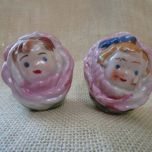 May include: Two porcelain salt and pepper shakers shaped like roses with painted faces. The girl shaker has a blue hat and the boy shaker has brown hair.