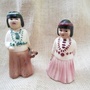 May include: Two ceramic figurines of Native American girls. One wears a green headband and necklace, a white top, and a brown skirt, with a small dog at her side. The other wears a red headband and a pink skirt, with a necklace.