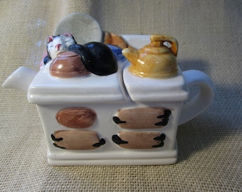 May include: White ceramic teapot shaped like a vintage stove with a black cat sleeping on top. The teapot has a brown and black painted design and a small yellow teapot lid.
