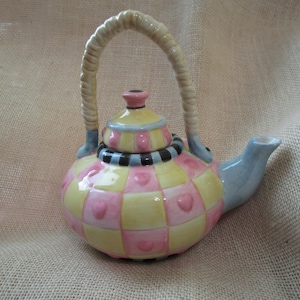 May include: A decorative ceramic teapot with a woven handle and a light blue spout. The teapot is painted with a checkerboard pattern of pink and yellow squares, each with a heart. The lid is yellow with pink accents.