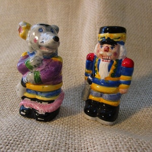 May include: A pair of ceramic figurines, a colorful mouse and a nutcracker. The mouse is wearing a purple, yellow, and blue outfit with a pink tail. The nutcracker is wearing a blue and yellow uniform with a red sash.
