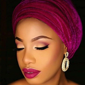 May include: A woman with dark skin is wearing a vibrant magenta velvet turban. Her eyes are closed and she is wearing dramatic eye makeup with a dark eyeliner and a light pink eyeshadow. Her lips are a deep magenta colour. She is wearing large gold hoop earrings with rhinestones.