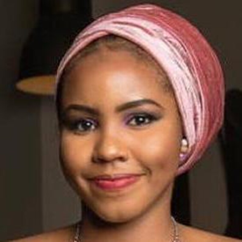 May include: A woman with dark brown skin is wearing a pink and rose-colored velvet turban. She is smiling and looking directly at the camera.