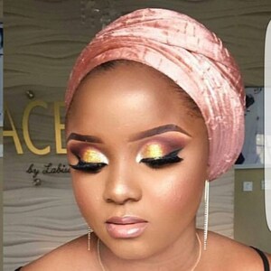 May include: A woman with dark skin is wearing a pink velvet turban. She has on dramatic eye makeup with gold and brown eyeshadow.