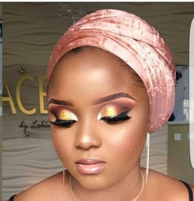 May include: A woman with dark skin is wearing a pink velvet turban. She has on dramatic eye makeup with gold and brown eyeshadow.