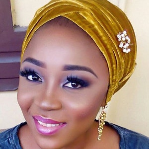 May include: A woman wearing a gold velvet turban with a pearl and rhinestone pin. She is wearing gold earrings and has a natural makeup look.