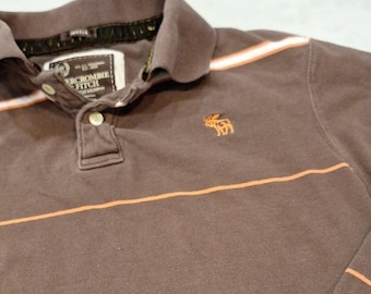 Puede incluir: Polo marrón con rayas naranjas y blancas. La camisa tiene cuello y tapeta de tres botones. El logotipo de Abercrombie & Fitch está bordado en el pecho.