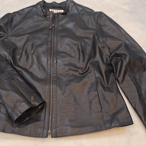 May include: A black leather jacket featuring a stand-up collar, a full-length front zipper, and long sleeves with zippered cuffs. The interior neck label displays the text A.M.I. The jacket has visible seam detailing across the front panels.