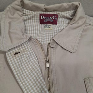 May include: Close-up of a tan jacket with a collar and a zipper. The jacket has a gingham lining and a label that reads "DENIM & Co." The jacket is made of a durable material and has a button closure.