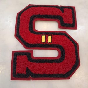 May include: A varsity-style letter S patch featuring a textured red center with a thick black border and a red felt backing. The letter includes two small, horizontal yellow embroidered bars positioned in the center of the letter's middle section.