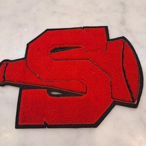 May include: A red chenille patch in the shape of the letter S, featuring a megaphone integrated into the design. The patch has a thick black border and a textured, fuzzy surface, creating a bold and distinct visual element for apparel.