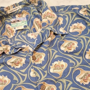 May include: Blue button-down shirt with a floral pattern in tan, pink, and green. The shirt has a collared neckline and a front pocket. The tag reads "Rayon Spooner". The shirt is made of cotton.