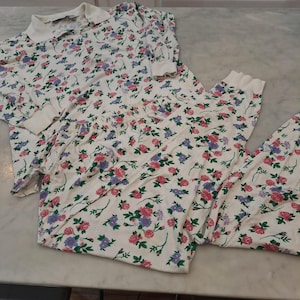 May include: A two-piece pajama set featuring a white background with a floral pattern of pink and purple roses and green leaves. The top has a collared neckline and long sleeves. The pants have an elastic waistband.