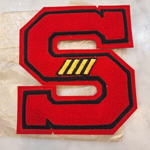 May include: A large, block-style letter S patch featuring a textured red surface with a thick black border. The center of the letter includes three diagonal yellow stripes. The patch is constructed with dense embroidery and has a bold, varsity-inspired design.