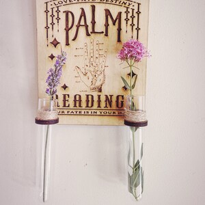Op de afbeelding: Een houten bord met de woorden "LOVE FATE DESTINY PALM READING YOUR FATE IS IN YOUR HANDS" en een hand met handlijnen. Twee glazen vazen met bloemen zijn aan het bord bevestigd.