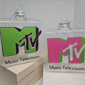 Puede incluir: Dos adornos cuadrados transparentes con el logotipo de MTV en verde y rosa. El logotipo está sobre un fondo verde y rosa, con las palabras "Music Television" impresas debajo. Cada adorno tiene una tapa plateada y una cuerda plateada para colgar.