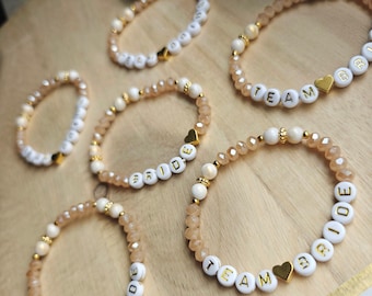 May include: Several beaded bracelets with white letter beads spelling "TEAM BRIDE" and "BRIDE". The bracelets feature peach-colored and white beads, gold heart charms, and gold accents. The bracelets are arranged on a wooden surface.