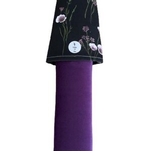 May include: A purple yoga mat carrier with a black floral patterned top. The top features white and purple flowers and green stems. A small white circular tag is visible on the top. The carrier has a black handle.