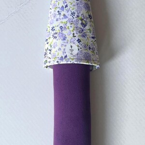 May include: A purple bolster pillow with a floral patterned cover. The cover is white with small purple and blue flowers. The pillow is approximately 12 inches long.