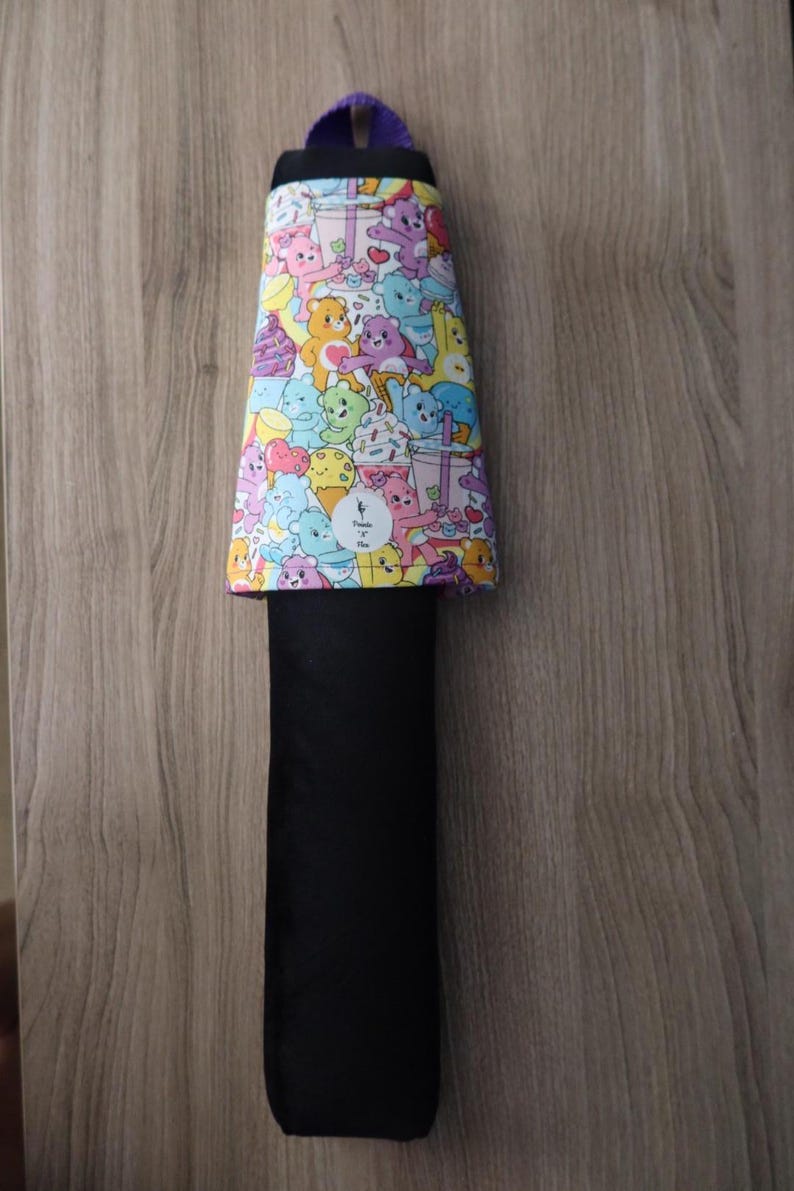 May include: A black and colorful Care Bears themed item. The top portion features a vibrant pattern of various Care Bears characters, ice cream, and other treats. The item has a black, rectangular body and a purple loop at the top.