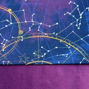 May include: A celestial-themed fabric featuring a deep blue background with gold and white constellations, sunbursts, and celestial map designs. The lower portion of the fabric is a solid, rich purple color. This fabric is suitable for various crafting projects.