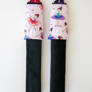 May include: Two car seat belt covers. Each has a black base and a light pink top with a repeating pattern of ballerinas, stars, and the text "Purple Pix". One has a pink strap, the other a purple strap.