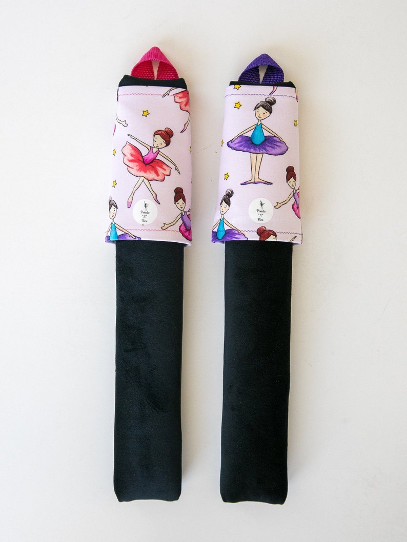 May include: Two car seat belt covers. Each has a black base and a light pink top with a repeating pattern of ballerinas, stars, and the text "Purple Pix". One has a pink strap, the other a purple strap.