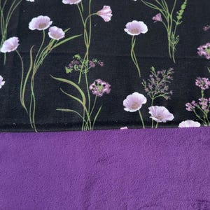 May include: A textile piece featuring a black fabric section with a printed pattern of light purple wildflowers and green stems, joined to a solid, textured purple fabric section. The floral print includes various small blossoms and sprigs.