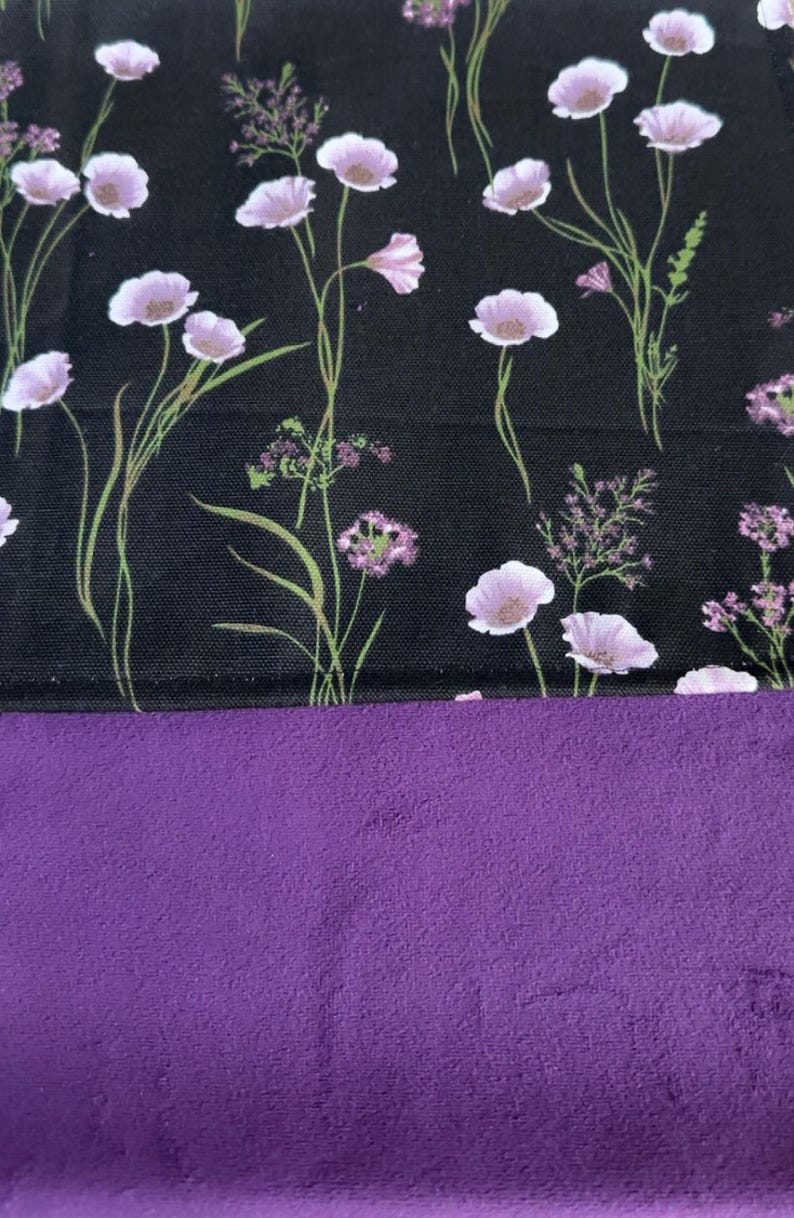 May include: A textile piece featuring a black fabric section with a printed pattern of light purple wildflowers and green stems, joined to a solid, textured purple fabric section. The floral print includes various small blossoms and sprigs.