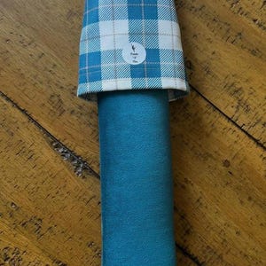May include: A teal-colored, cylindrical item with a plaid top in white and teal. The item has a loop at the top and a small circular logo. The item is likely a sleeve or cover for a cylindrical object.