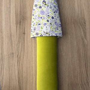 May include: A green door stop with a floral patterned top in white, purple, and green. The door stop has a purple loop at the top for hanging. The floral pattern includes small flowers and leaves. The door stop is on a wooden surface.
