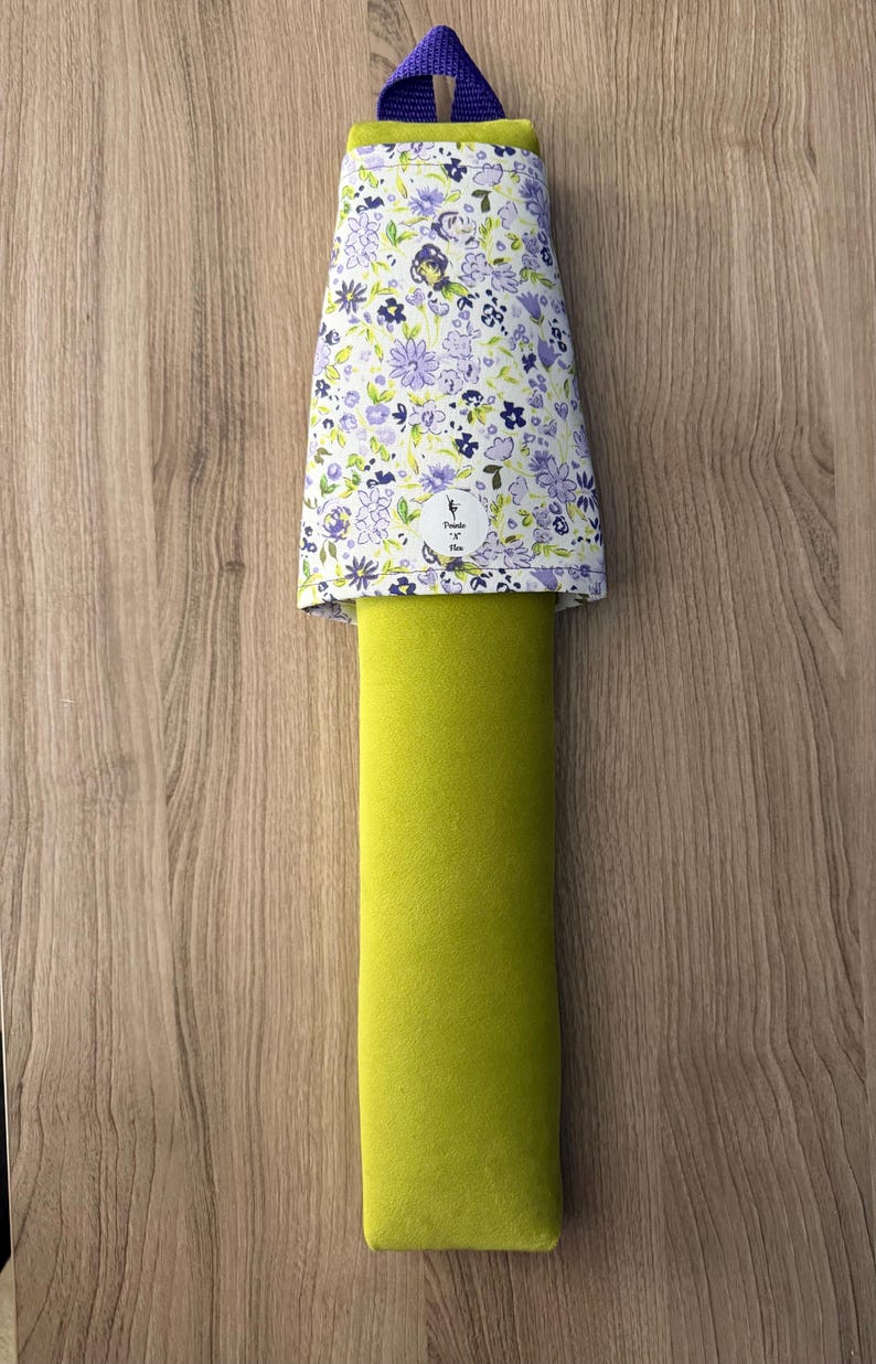 May include: A green door stop with a floral patterned top in white, purple, and green. The door stop has a purple loop at the top for hanging. The floral pattern includes small flowers and leaves. The door stop is on a wooden surface.