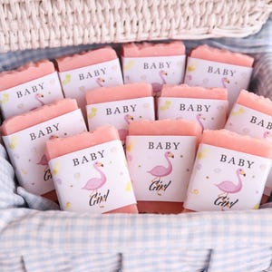 May include: Pink baby soaps with flamingo graphics and the word "Baby" on the labels, arranged in a woven basket with a blue and white checkered lining. The soaps are a light pink color.