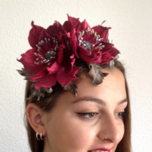 May include: A burgundy floral headband with two large fabric flowers and silver accents. The headband is worn by a person with long brown hair.