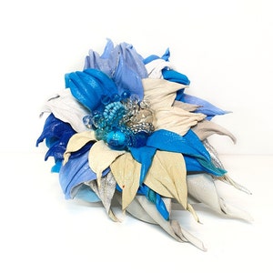 Könnte beinhalten: Handgefertigte Lederblume in Blau-, Silber- und Beigetönen. Die Blume hat einen Cluster aus blauen Perlen und klaren Kristallen in der Mitte. Die Blütenblätter sind aufwendig geschnitten und geformt, wodurch ein mehrschichtiger Effekt entsteht.