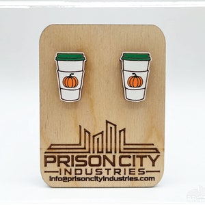 May include: A pair of wooden earrings shaped like a pumpkin spice latte. The earrings are white with a green lid and a small orange pumpkin on the side. The earrings are on a wooden display card with the text "Prison City Industries" and a website address.