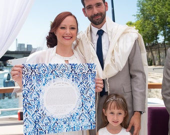 May include: A couple poses for a wedding photo with a blue and white ketubah, a traditional Jewish marriage contract, in front of a white backdrop. A young girl in a white dress stands in front of them.