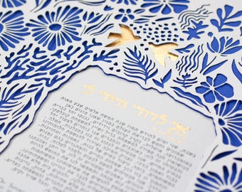 May include: A white laser-cut paper frame with intricate floral and leaf patterns layered over a solid blue background. Two gold-foiled birds are positioned at the top centre. The central area features Hebrew text printed in gold foil.