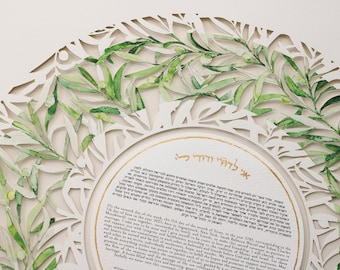 May include: A white circular ketubah with a gold border and a watercolor design of olive branches. The text in Hebrew reads "Blessed are you, Lord our God, King of the Universe, who has sanctified us with His commandments and commanded us to be bound in the covenant of marriage."