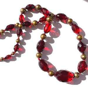 May include: A necklace featuring faceted, deep red beads interspersed with smaller gold-colored beads. The necklace is strung on a red cord and has a gold-colored clasp. The beads have a faceted cut, reflecting light.