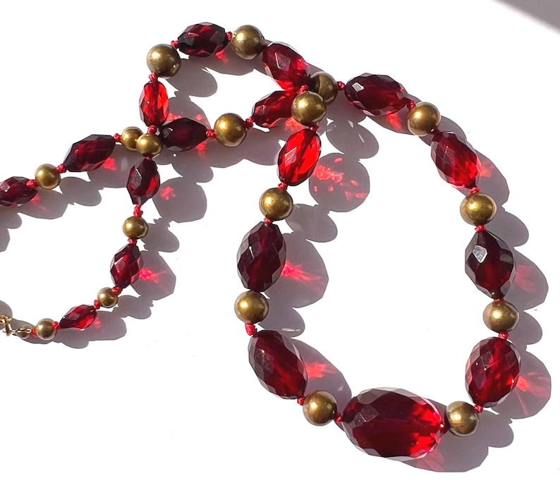 May include: A necklace featuring faceted, deep red beads interspersed with smaller gold-colored beads. The necklace is strung on a red cord and has a gold-colored clasp. The beads have a faceted cut, reflecting light.