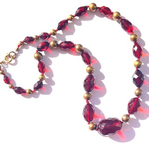 May include: A vintage necklace featuring faceted, deep red glass beads and small gold-toned beads. The necklace has a lobster clasp closure. The beads are strung on a red cord. The necklace is displayed on a white surface.